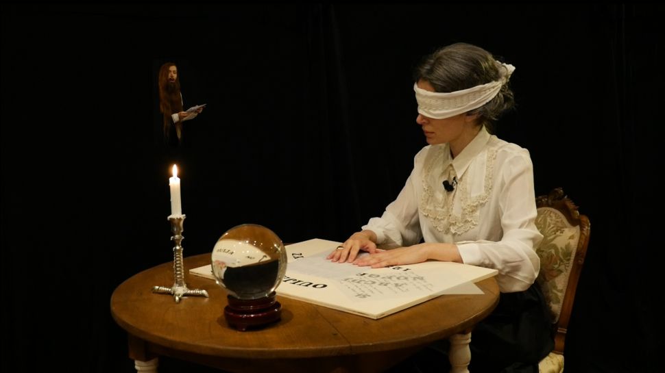 A staged performance scene showing a blindfolded figure seated at a table with a candle, crystal ball and open book in a darkened setting.