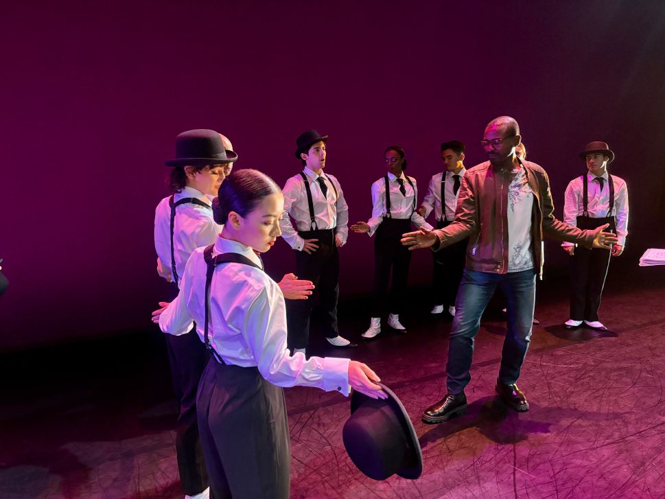 A choreographer stands on stage giving instructions to a group of dancers dressed in white shirts, suspenders, and bowler hats during a rehearsal under deep purple lighting.