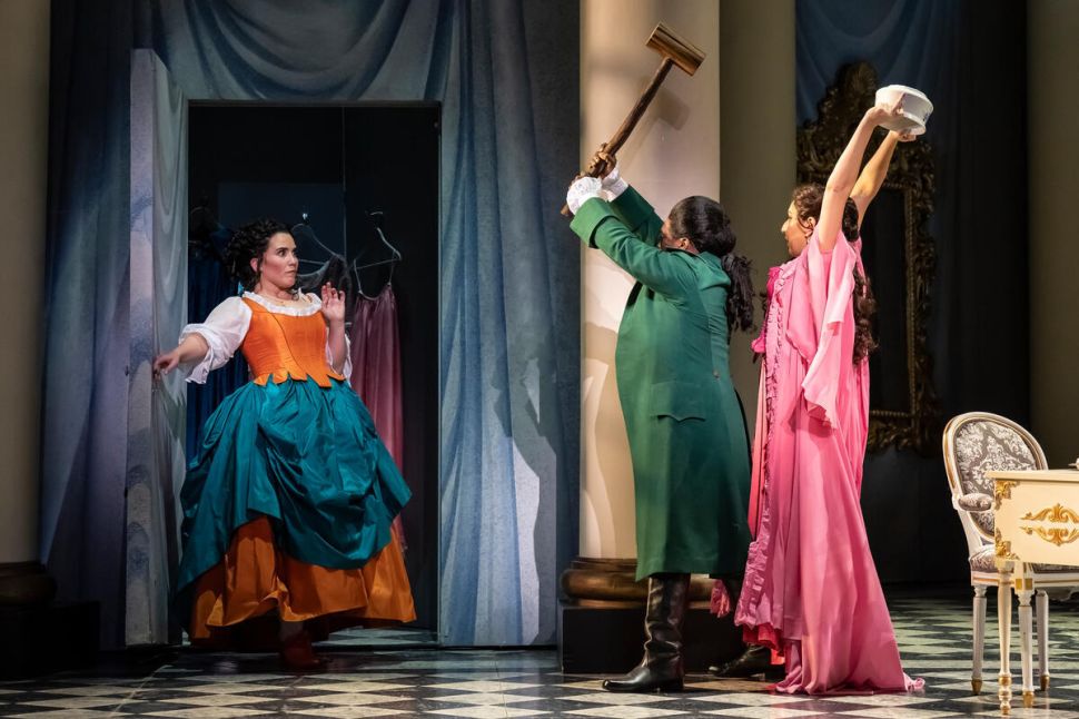 A scene from Le Nozze di Figaro showing three performers: a woman in a colorful dress with a corset and full skirt holding her hand up in surprise, a man in a green coat with boots holding a large wooden mallet as though about to strike, and a woman in a pink gown raising her arm, standing next to a white chair, on a tiled floor with draped curtains and a wardrobe in the background.