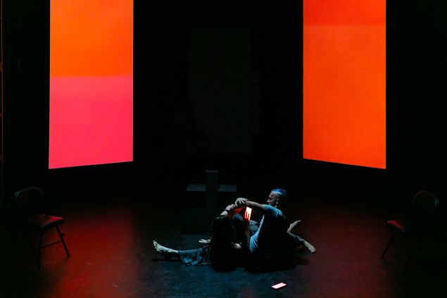 Two performers sit on a dark stage illuminated by large glowing red and orange screens, creating an abstract digital environment around them.