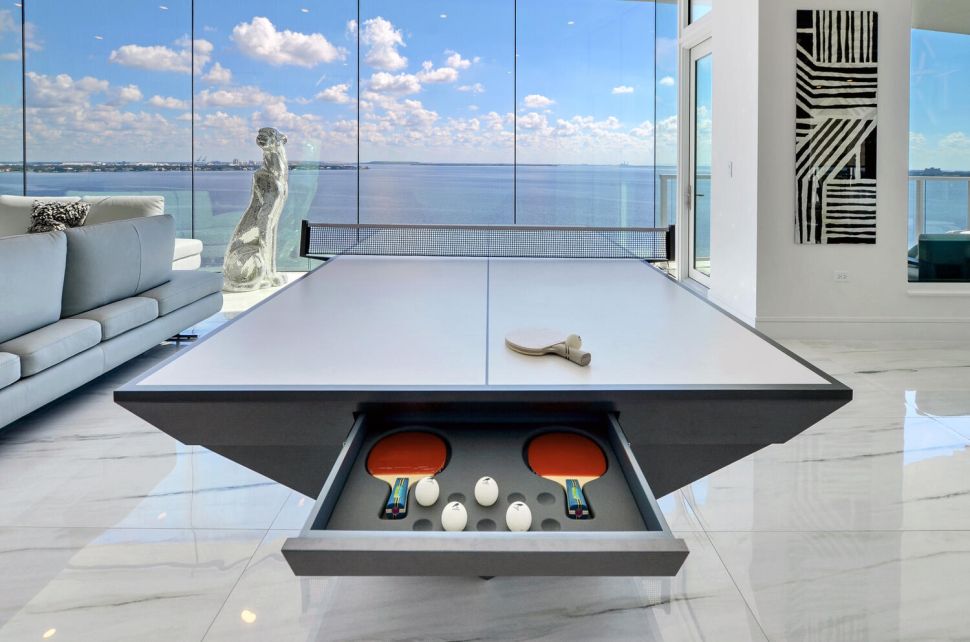 Modern luxury ping pong table with hidden paddle drawer in a high-rise waterfront residence, featuring panoramic ocean views and contemporary art.