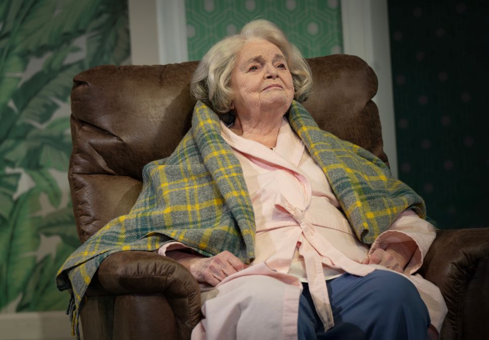 A photograph shows June Squibb sitting alone in a recliner wrapped in a plaid blanket, looking upward with a calm but distant expression in a scene from Marjorie Prime.
