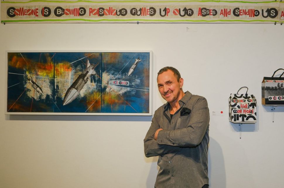 A man in a dark gray button down shirt stands smiling with his arms folded in front of a display of artworks
