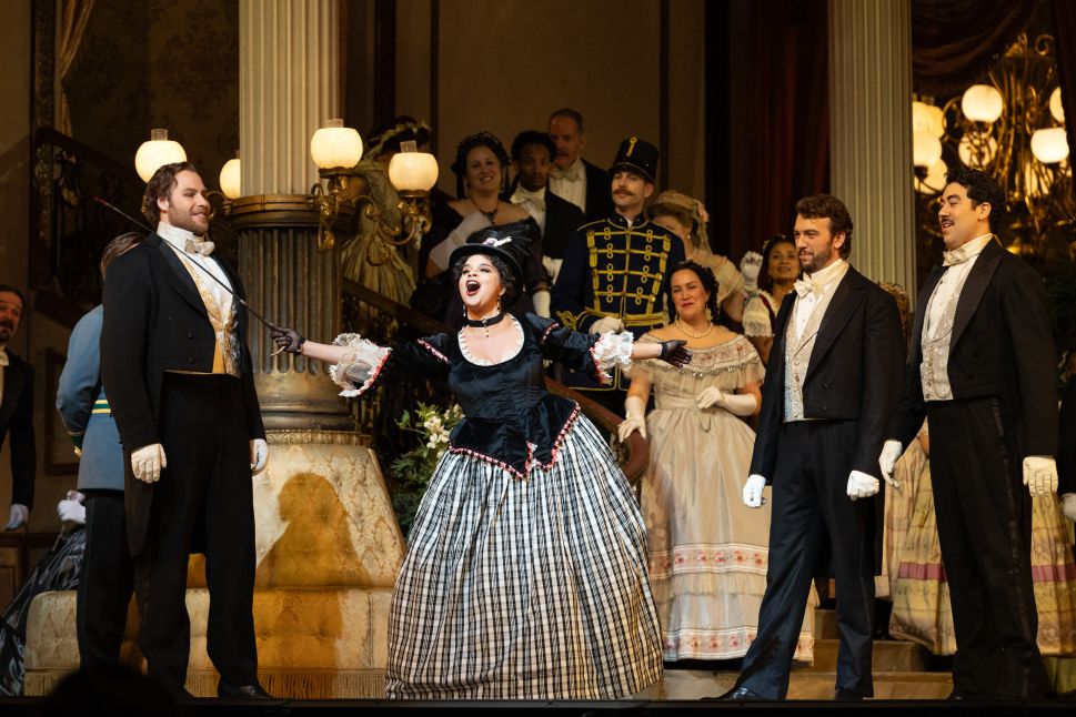 A female singer in a large black-and-white gown sings expressively at the center of a crowded ballroom scene as male and female cast members look on, capturing an ensemble moment from the Metropolitan Opera’s Arabella.