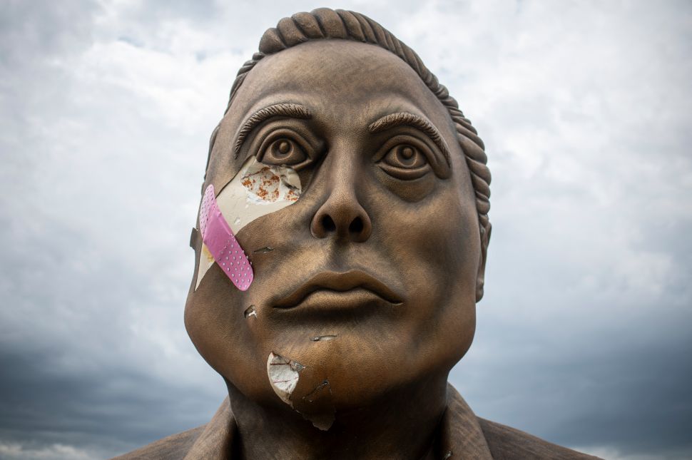 Close-up of a weathered bronze statue with visible cracks, peeling paint, and a pink bandage on its cheek, set against a cloudy sky.