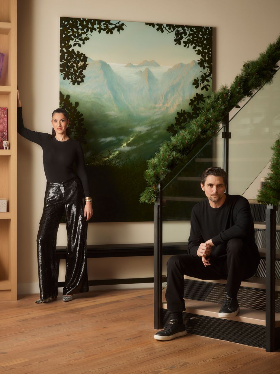 A woman stands beside a large landscape painting while a man sits on a staircase in a modern interior decorated with holiday greenery.