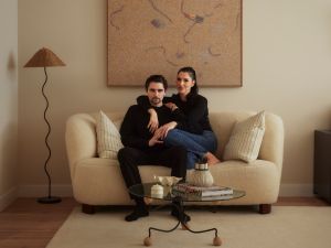 A couple dressed in black sit closely together on a cream-colored sofa beneath an abstract painting in a softly lit living room.