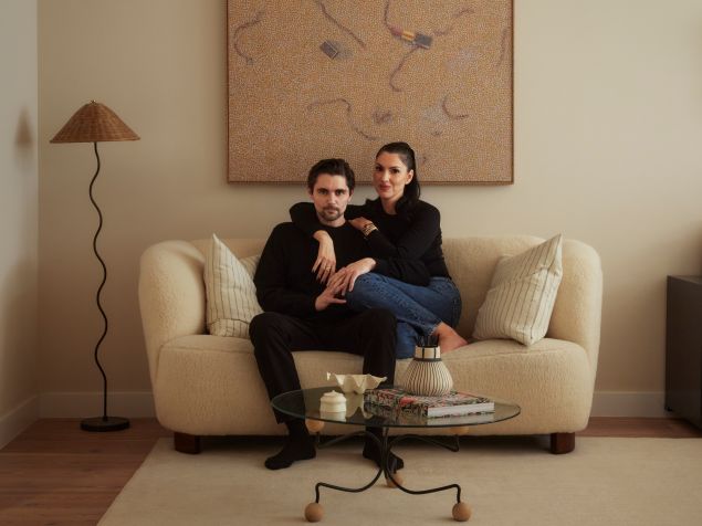 A couple dressed in black sit closely together on a cream-colored sofa beneath an abstract painting in a softly lit living room.