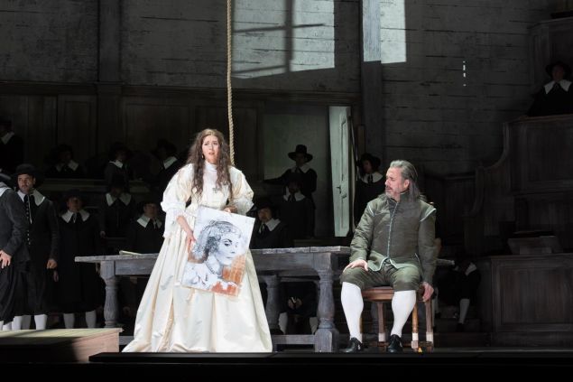 A woman in a white gown holds up a rough sketch of a face during a tense scene on stage while a man sits beside her and a chorus watches from the background.