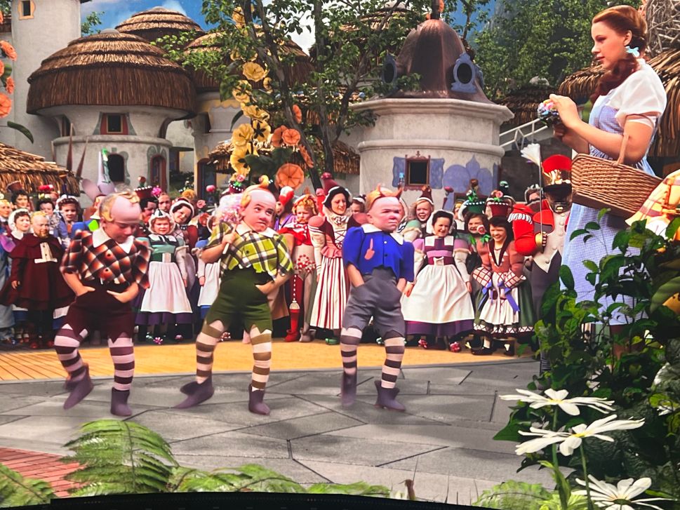 A large crowd of brightly dressed performers stands in a colorful village scene while three small dancers perform in front of a girl holding a basket, evoking the Munchkinland sequence from The Wizard of Oz.