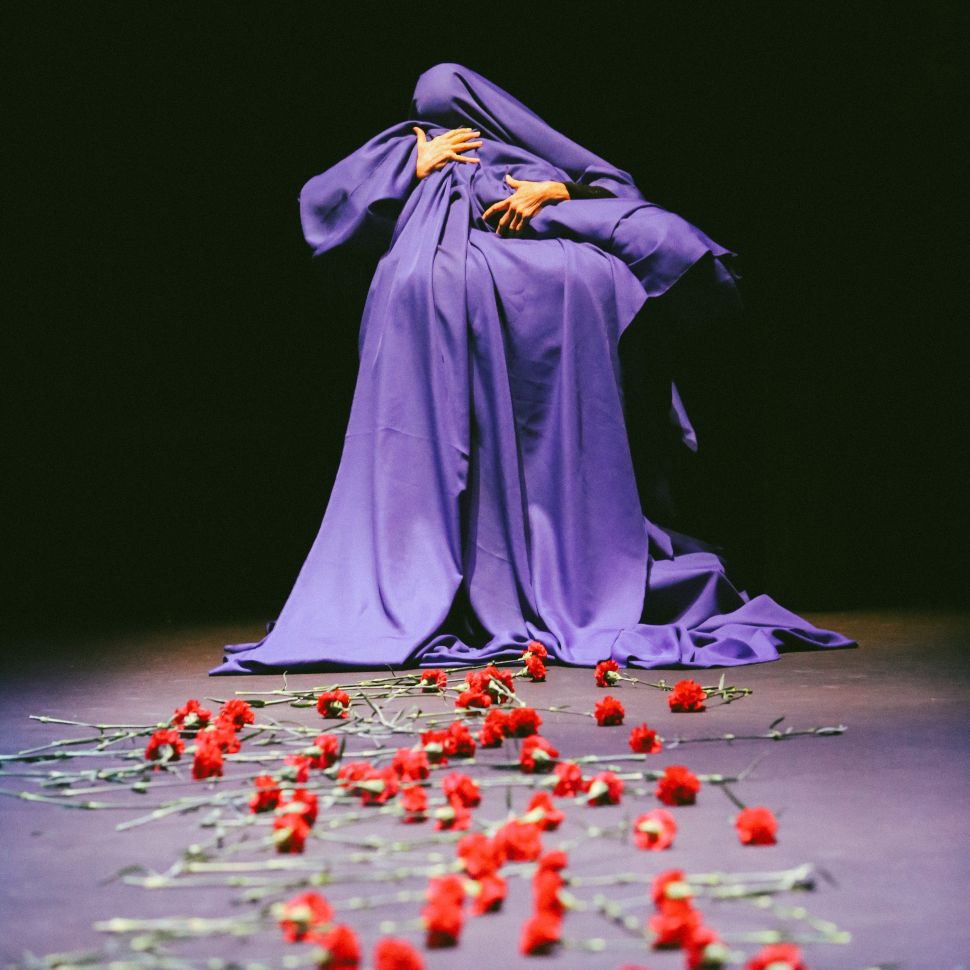 A figure shrouded in blue holds a body-shaped bundle against a black background. They are framed at a distance. Red carnations with green stems trail from the feet of the figure to the foreground of the picture before blurring out of focus.