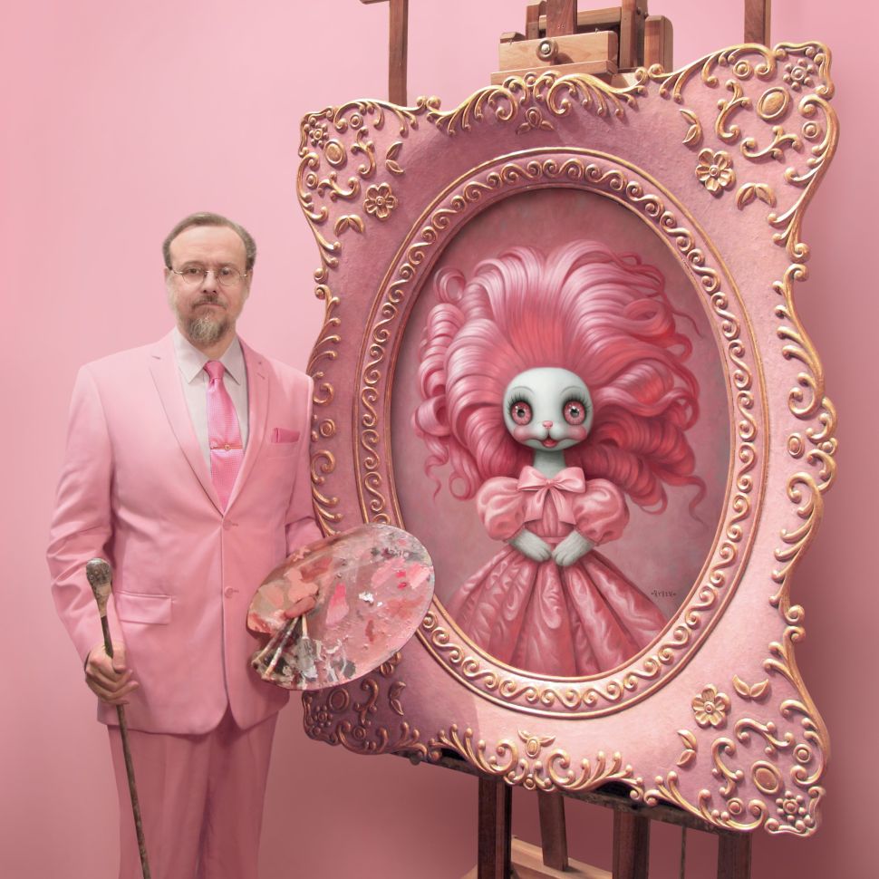 Man in a pastel pink suit stands beside an ornate easel, holding a paint palette and brush. On the canvas is a surreal, wide-eyed doll with voluminous pink hair and a bow, framed in gilded pink. The studio background is entirely pink.