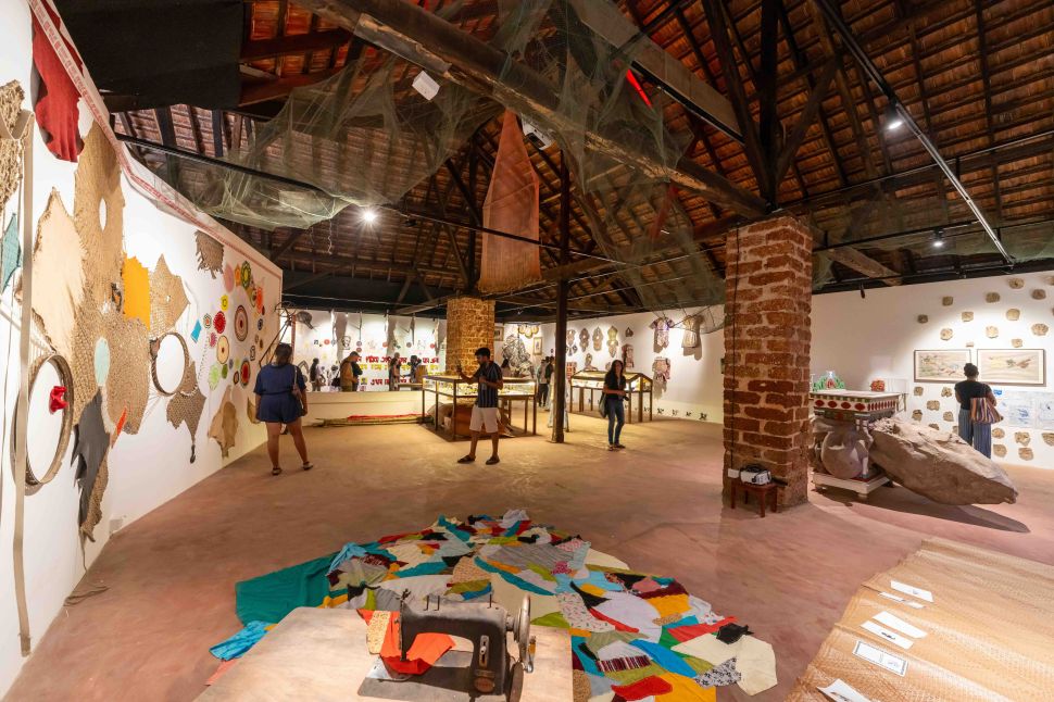 A gallery interior filled with textile works, vitrines, and hanging materials is viewed from a wide central aisle.