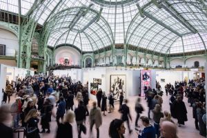 A wide view of a historic glass-roofed exhibition hall filled with art fair booths and crowds circulating across multiple levels.