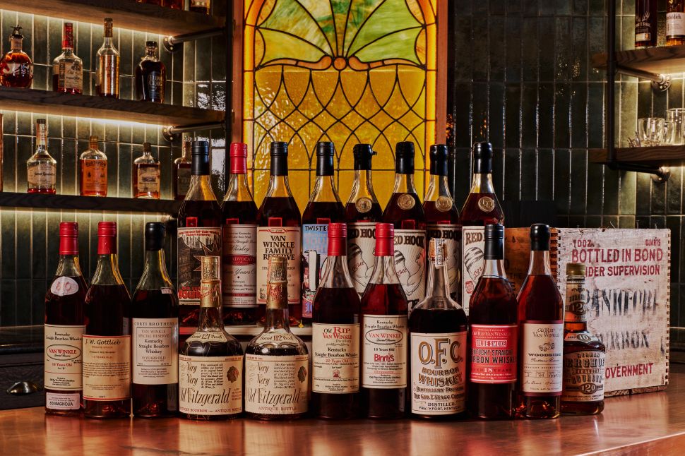 A curated lineup of rare American whiskey bottles displayed on a wooden bar, featuring vintage bourbons and rye labels arranged against backlit shelves and stained glass.
